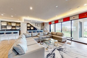 A modern living room with a sectional sofa, a glass coffee table, and a view of a green landscape through large windows. - The Epic Portal Mansion (Kissimmee)