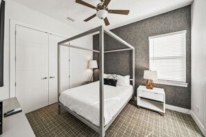 A bedroom with a four-poster bed, a bedside table with a lamp, a window with blinds, and a ceiling fan. - Modern Villa With Games Room at Reunion (Kissimmee)
