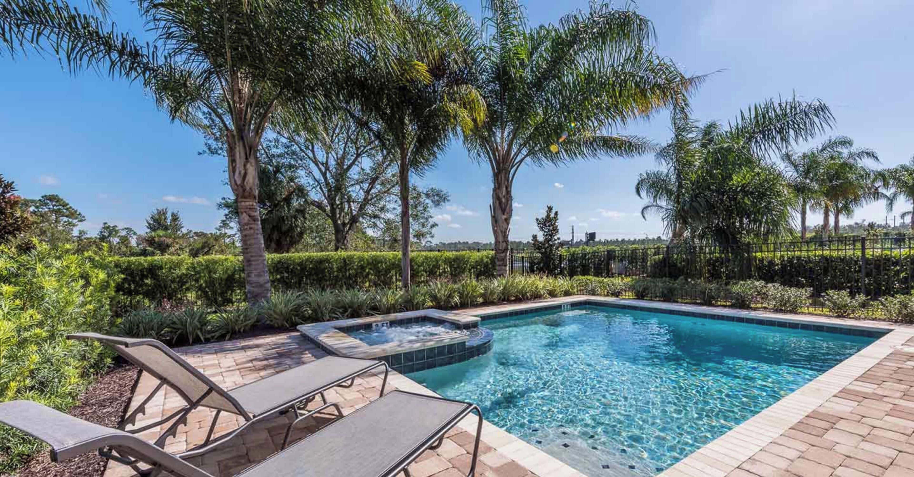 A swimming pool with lounge chairs and a hot tub, surrounded by lush greenery and a clear sky.