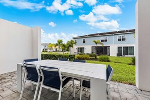 Une terrasse extérieure avec une table et des chaises blanches, un ciel bleu dégagé et un immeuble d’habitation en arrière-plan.