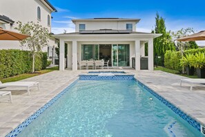 A swimming pool with a tiled edge, a covered patio area with outdoor furniture, and a building with large windows and a flat roof. - The Villa Tranquility With Private Pool (Kissimmee)