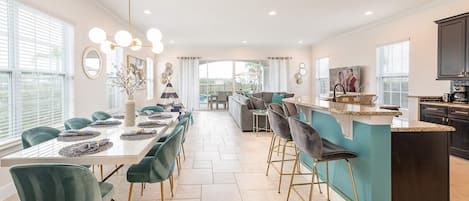 A spacious dining area with a long table and chairs, a kitchen with dark cabinets, and a view through the windows.