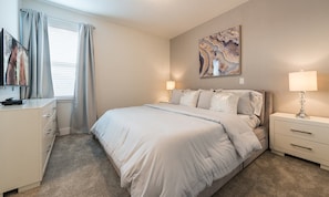 Ein Schlafzimmer mit Bett, Nachttischen, einer Kommode, einem Bild und einem Fenster mit Vorhängen.