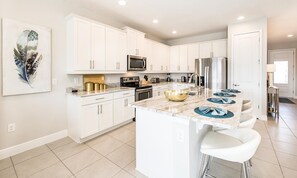 A modern kitchen with white cabinetry, a central island with bar stools, stainless steel appliances, and a decorative bowl. - Spacious Home With South Facing Pool in Encore (Kissimmee)