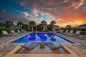 A rectangular swimming pool with lounge chairs and a small table in the center, surrounded by palm trees and buildings. - Spacious Home With South Facing Pool in Encore (Kissimmee)