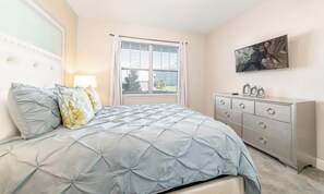 Une chambre à coucher avec un lit, une commode, une fenêtre avec des stores et un tableau accroché au mur.