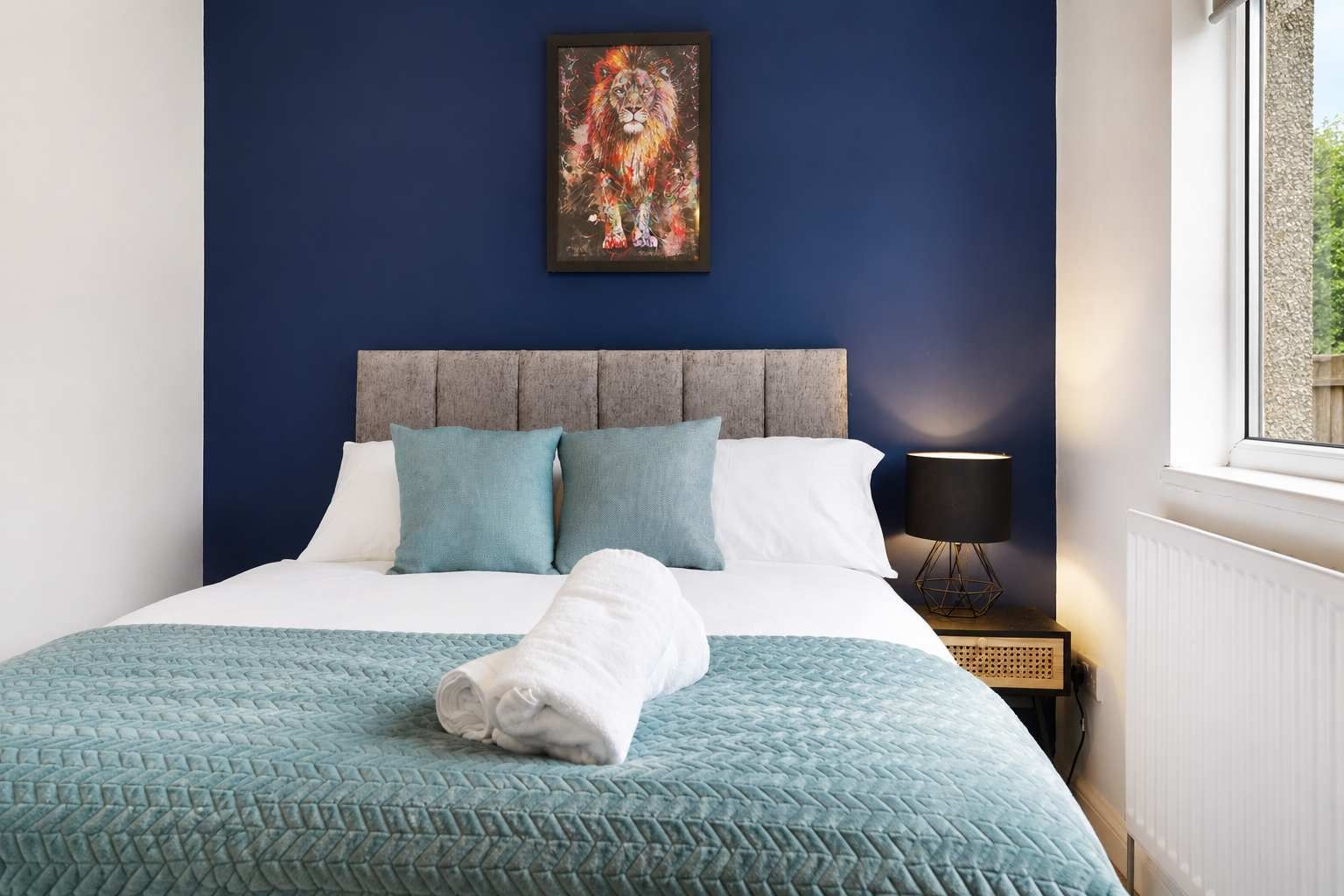 A neatly made bed with a blue quilt, pillows, and a white towel, a painting on the wall, and a bedside lamp.