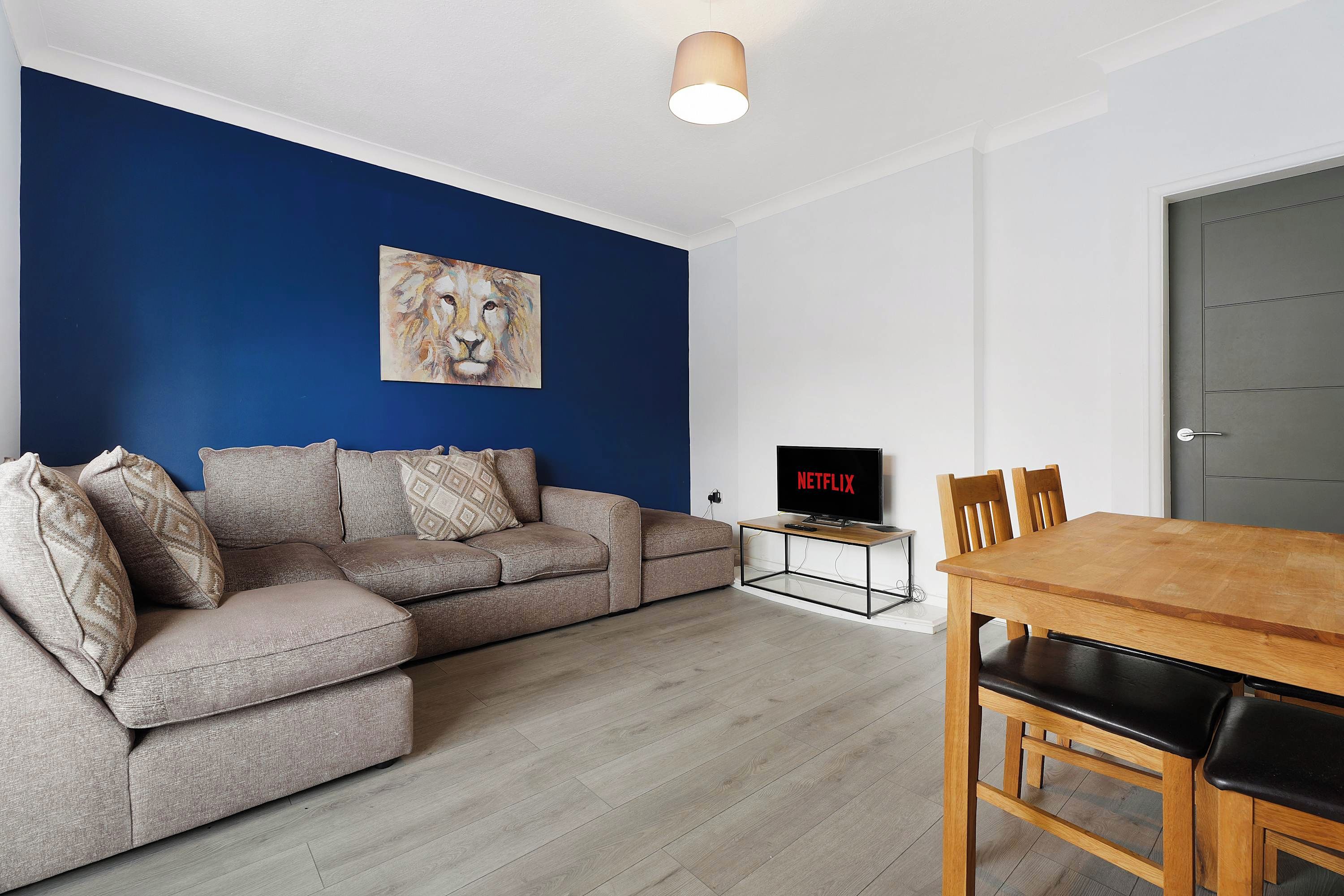 Un salon moderne avec un mur d’accent bleu, un canapé gris, une table à manger en bois et un téléviseur à écran plat affichant le logo « NETFLIX ».