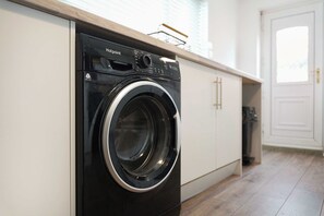A modern, compact washing machine in a clean, minimalist kitchen with white cabinetry and a wooden countertop. - Chic 4BR Contractor Stay for 7+WiFi | Stay With Us (Stockton-on-Tees)
