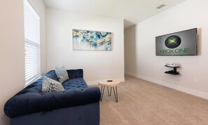 A living room with a dark blue sofa, a small round table, a wall-mounted Xbox One console, and a piece of abstract art. - Stylish Villa With Unreal Themed Bedroom at Reunion (Kissimmee)