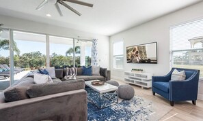 A modern living room with a large sofa, a coffee table, and a blue armchair. There is a TV mounted on the wall and a large window providing a view of the outdoors. - Stylish Villa With Unreal Themed Bedroom at Reunion (Kissimmee)