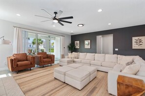 Ein modernes Wohnzimmer mit einem Ecksofa, zwei Sesseln, einem Couchtisch, einem Deckenventilator und einem großen Fenster mit Blick ins Freie.