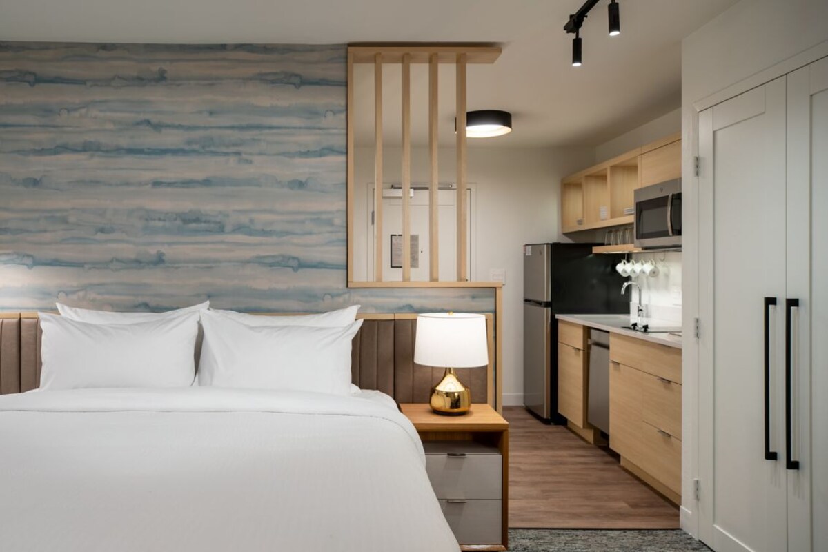 A hotel room with a large bed, a bedside table with a lamp, a wooden headboard, a wall with a blue-gray pattern, a kitchen area with wooden cabinets, and a microwave.