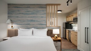 A hotel room with a large bed, a bedside table with a lamp, a wooden headboard, a wall with a blue-gray pattern, a kitchen area with wooden cabinets, and a microwave. - Fairfield Inn & TownePlace Suites  (La Porte)