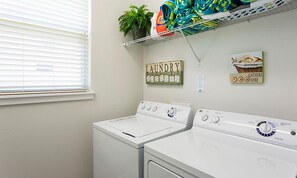 A laundry area with two front-loading washing machines, a shelf with folded towels, a potted plant, and a sign indicating a laundry gathering area. - Stylish Home With Themed Bedroom at Reunion (Kissimmee)