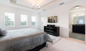 A bedroom with a large bed, a dresser, a TV, and a bathroom visible through an archway. - Ultra-modern Villa On Golf Course at Reunion (Kissimmee)