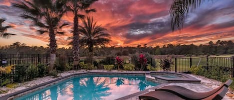 A swimming pool with lounge chairs, surrounded by palm trees and a fence, with a sunset in the background.