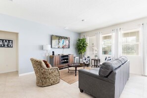 Ein modernes Wohnzimmer mit grauem Sofa, einem gemusterten Sessel und einem Couchtisch. Ein Fernseher steht auf einer Holzkommode, daneben ein Blumentopf und ein Bild an der Wand.