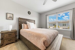 A bedroom with a bed, bedside tables, a clock, a window with blinds, and a ceiling fan. - The Serenity Townhome (Kissimmee)
