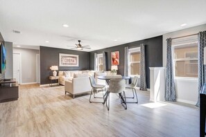 A modern living room with a sofa, dining table, and chairs, a ceiling fan, and large windows with curtains. - Orlando 6BR Villa With Private Pool at Windsor Westside (Kissimmee)