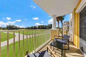 A balcony with a view of a green lawn and a pathway, a chair, a table, and a building with yellow walls. - The Golf Vista Townhome at Reunion (Kissimmee)