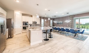 A modern kitchen with stainless steel appliances, a marble island, and a dining area with blue chairs and a pool view. - Gleaming Villa With Themed Bedrooms at Encore (Kissimmee)