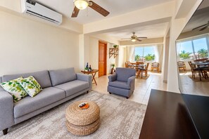 A living room with a grey sofa, a blue armchair, a woven ottoman, and a dining area with wooden furniture. - Hard Rock Central Suite,  Balcony, Parking (San Felipe de Puerto Plta)