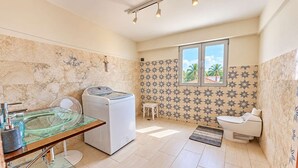 Un baño con lavadora, un lavabo de vidrio, un inodoro y una ventana con vistas a palmeras.