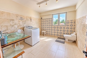 Ein Badezimmer mit Waschmaschine, einem Glastischwaschbecken, Toilette und einem Fenster mit Blick auf Palmen.