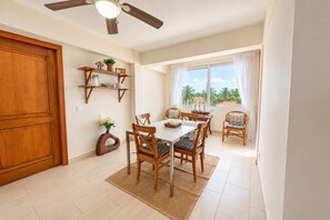 Ein Essbereich mit Tisch, Stühlen, Deckenventilator und Blick auf Palmen durch das Fenster.