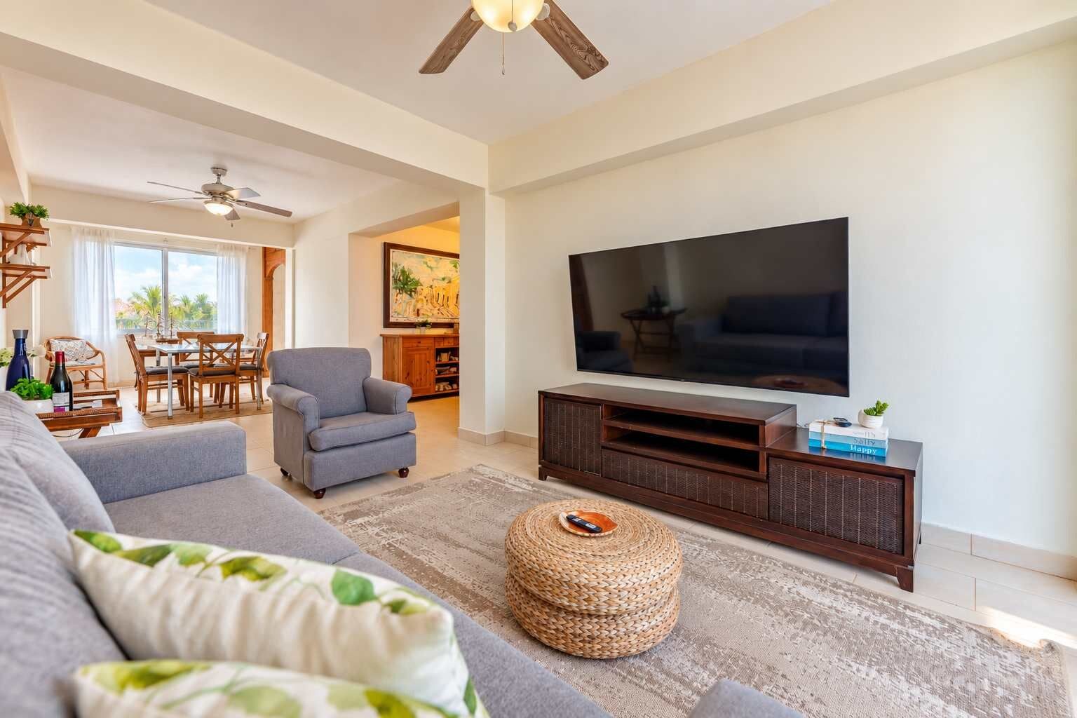 Ein Wohnzimmer mit grauem Sofa, einem Fernseher auf einem Holztisch, einem gewebten Hocker und einem Essbereich im Hintergrund.