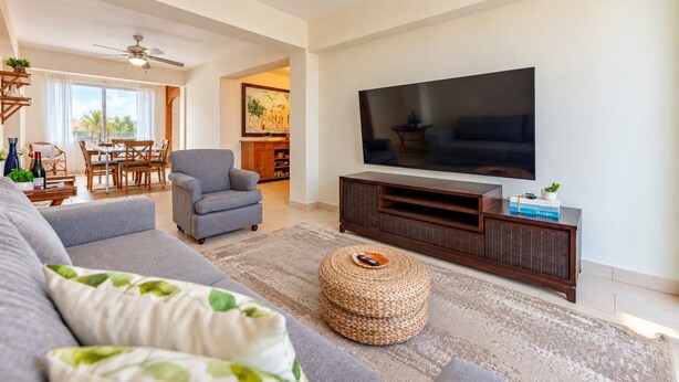 Una sala de estar con un sofá gris, un televisor sobre un mueble de madera, un otoman tejido y un área de comedor al fondo.