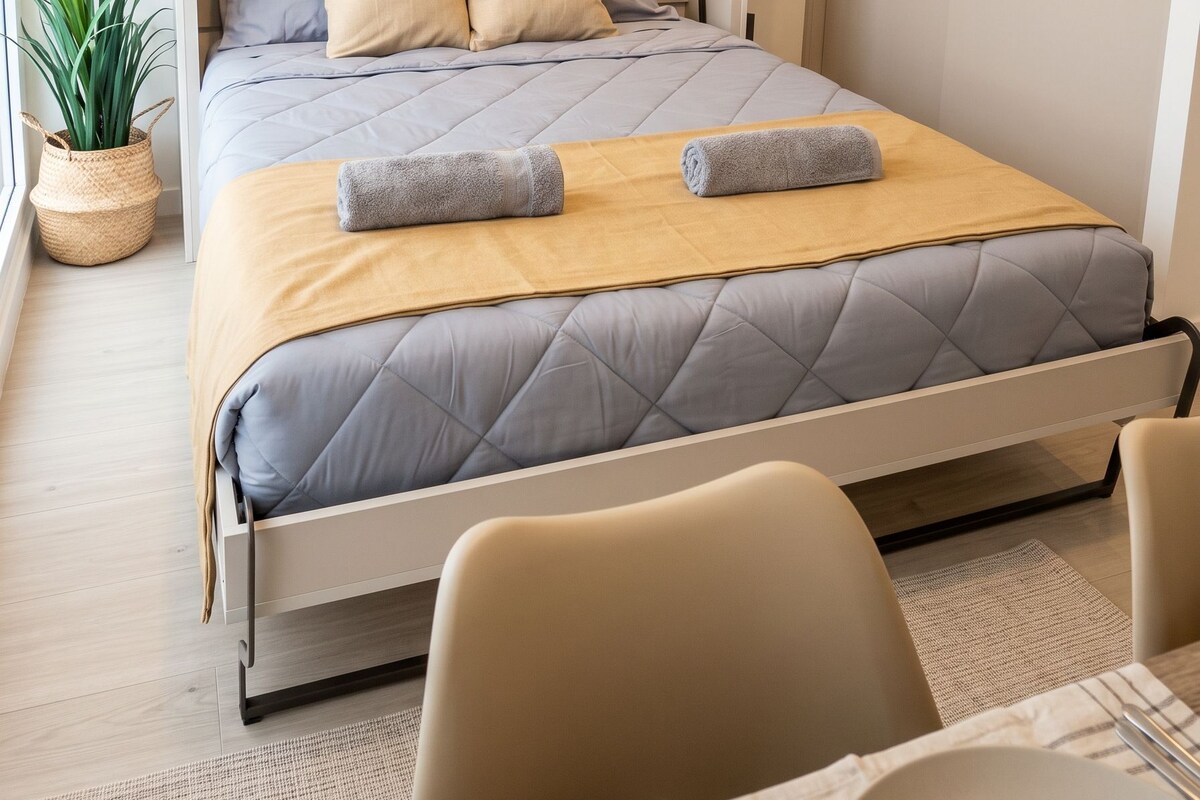 A modern bedroom with a bed, pillows, a bedside table, a plant, and a chair.