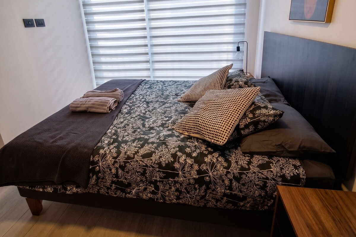 A bedroom with a bed, a window with blinds, a painting on the wall, and a bedside table.