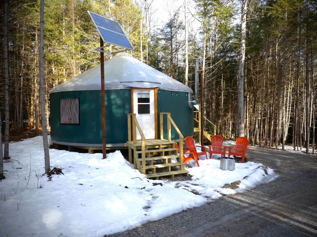 A yurt with a door, a small window, and a solar panel on top. There are orange chairs outside and a wooden ramp leading to the door.
