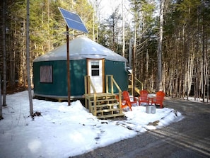 A yurt with a door, a small window, and a solar panel on top. There are orange chairs outside and a wooden ramp leading to the door.