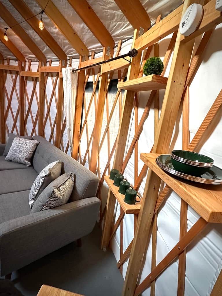 Ein Zimmer mit Holzbalkendecke, einem grauen Sofa mit gemusterten Kissen und einem Regal mit einer grünen Schüssel und Tassen.