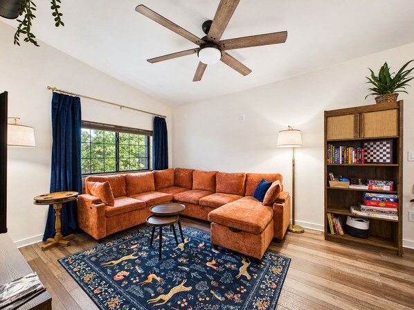 A living room with a sectional sofa, a wooden floor, a ceiling fan, a bookshelf, and a window with curtains.
