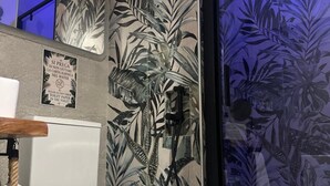 Et badeværelse med et moderne toilet, en bruseniche med et tropisk bladmotiv, og et skilt, der instruerer om ikke at skylle toiletpapir i toilettet.
