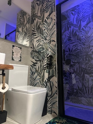 Un bagno con un water moderno, un box doccia con motivo a foglia tropicale e un cartello che indica di non gettare la carta igienica nel water.
