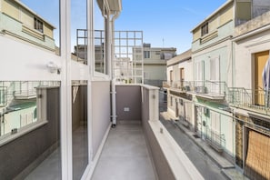 A narrow corridor with a glass door leading to a balcony, surrounded by buildings with balconies and railings. - 'Moonlight Jungle' Apartment with Balcony, Wi-Fi and Air Conditioning (Avola)