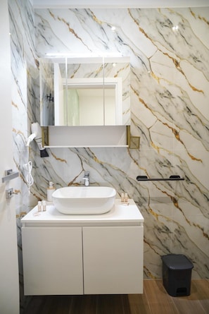 Un bagno con rivestimento in marmo, un mobile lavabo bianco con lavandino, uno specchio sopra il mobile e uno sgabello nero.