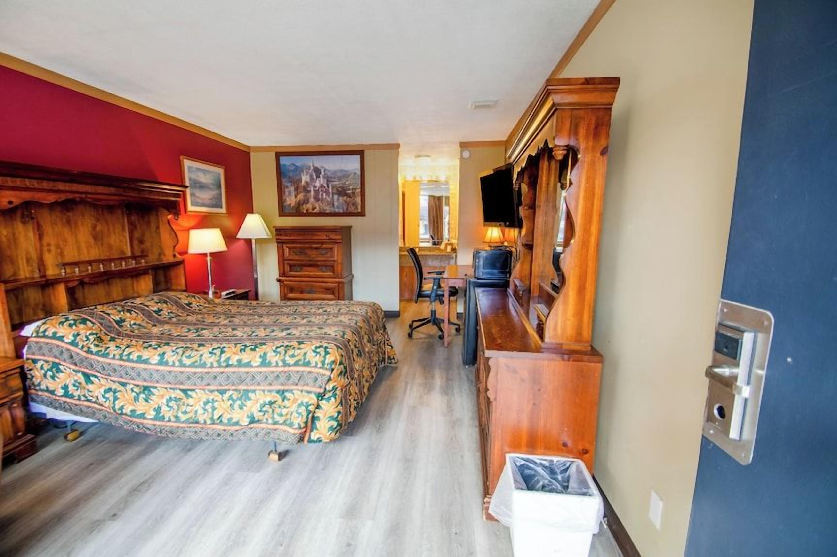 A hotel room with a wooden bed, a desk, a chair, a television, and a trash bin.