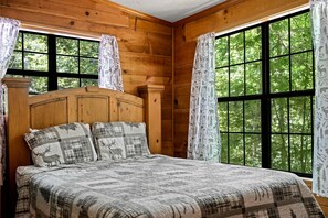 Ein Zimmer in einer Holzhütte mit einem Bett, Vorhängen und großen Fenstern.
