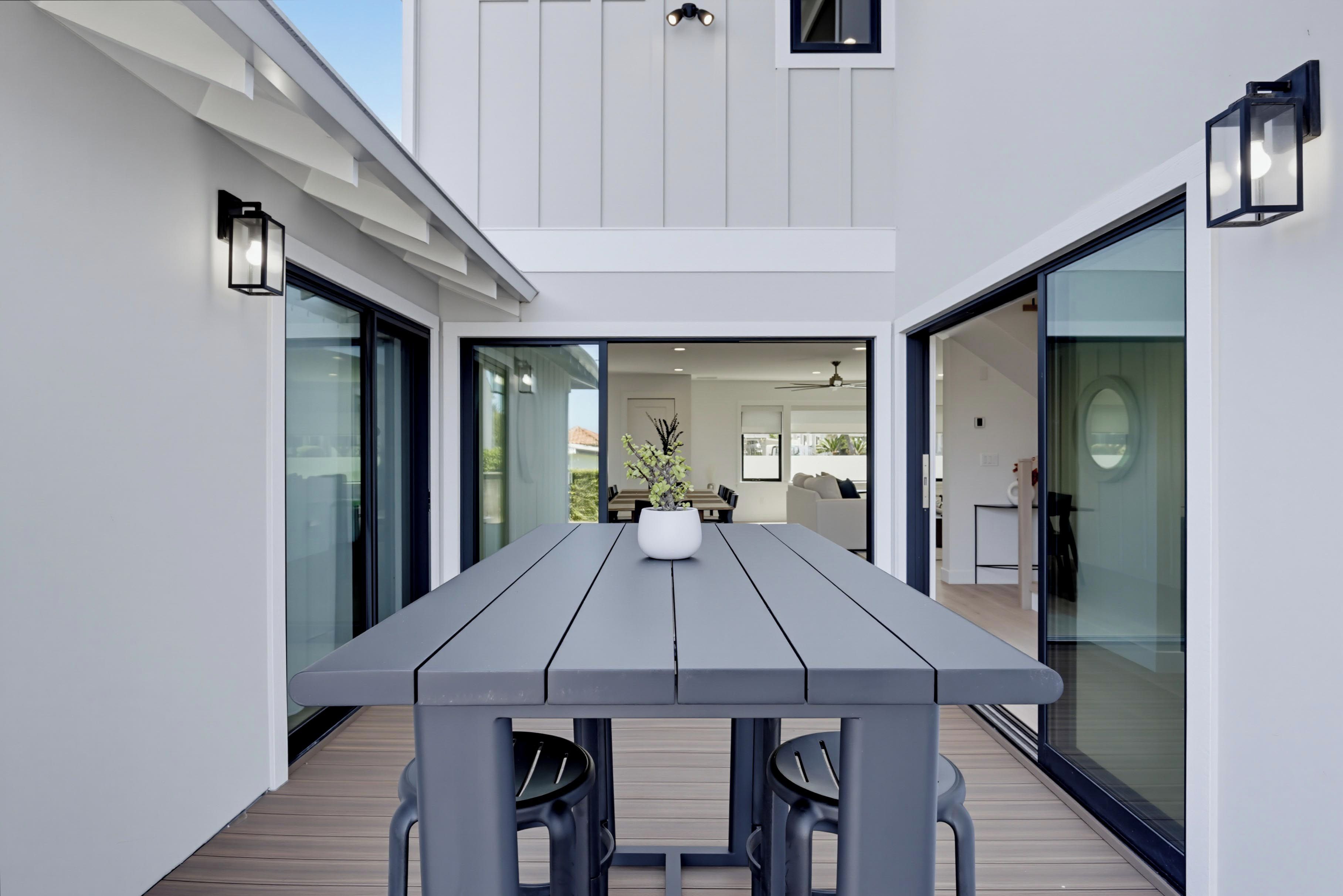 A modern outdoor dining area with a wooden table and chairs, a potted plant, and a view into the interior of the house.