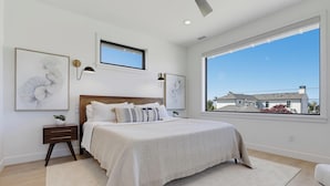 A bedroom with a large window, a bed with multiple pillows, a bedside table, and wall-mounted artwork. - Modern Coastal Home in Carlsbad (Carlsbad)