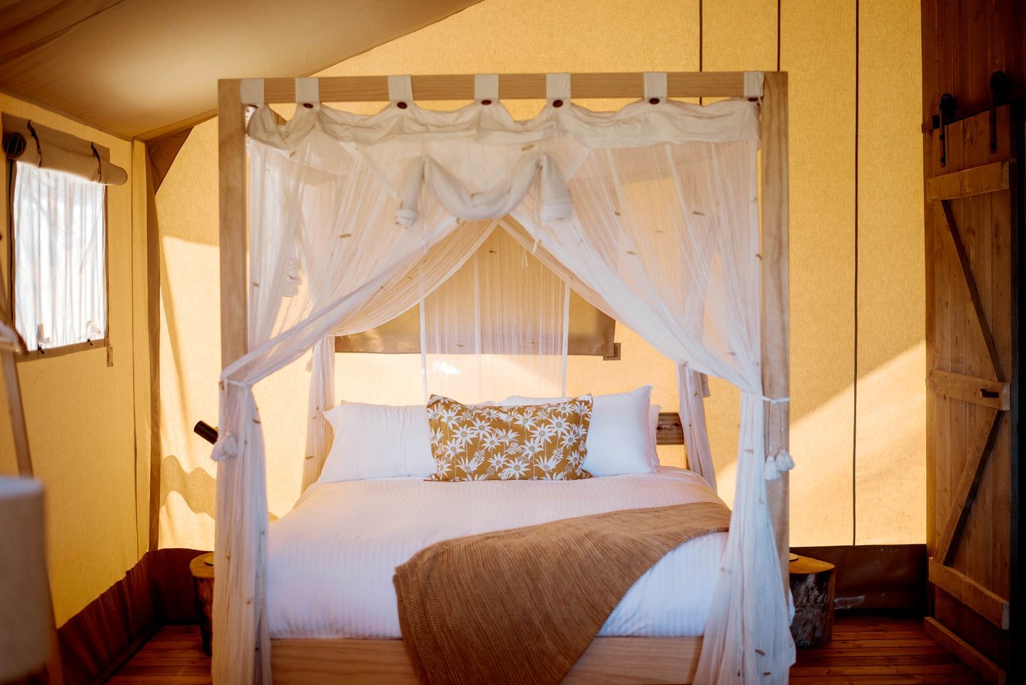 Un lit à baldaquin à quatre poteaux, agrémenté de draps blancs et d’un coussin à motifs floraux, placé dans une pièce aux murs en bois et dotée d’une fenêtre.