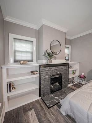 Une chambre avec une cheminée, un lit, une fenêtre avec des stores et un miroir au mur.
