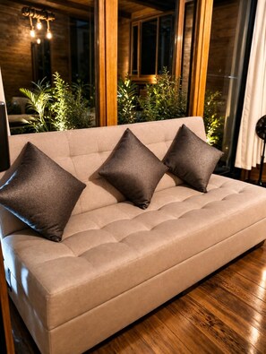 Un sofá con tres almohadas en una habitación con suelo de madera y ventanas amplias.