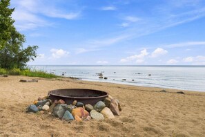 Ein Strand mit einem Feuerkorb, umgeben von Felsen, bewölktem Himmel und ruhigem Wasser.